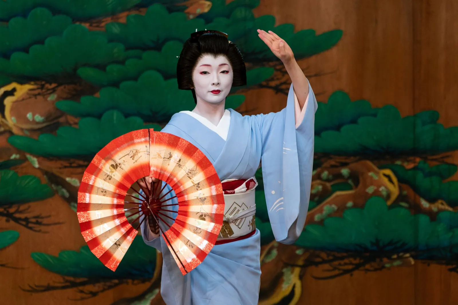 Geiko ve světle modrém kimonu tančí na jevišti před malovanou borovicovou stěnou, jednu ruku zvednutou a v druhé otevřený červenobílý vějíř
