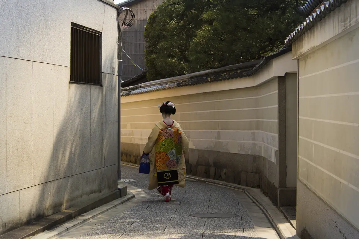 Eine Maiko in einem gelb gemusterten Kimono geht eine schmale Gasse zwischen Putzwänden in Kamishichiken entlang, ihr langes Obi hängt ihr über den Rücken