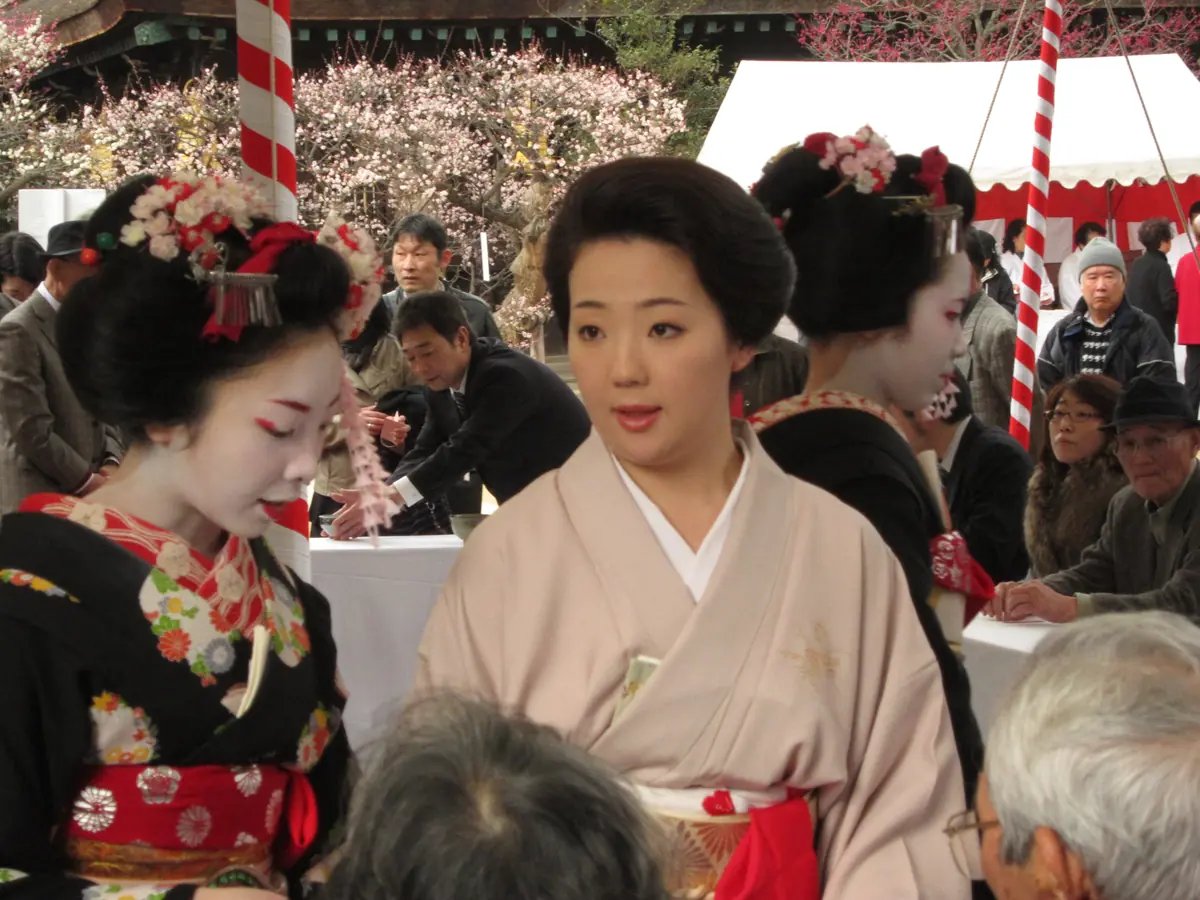 Eine Geiko in einem blassrosa Kimono steht zwischen zwei Maiko mit aufwendigen Blumenhaarschmuck beim Pflaumenfest im Kitano Tenmangū, dahinter sitzen Besucher