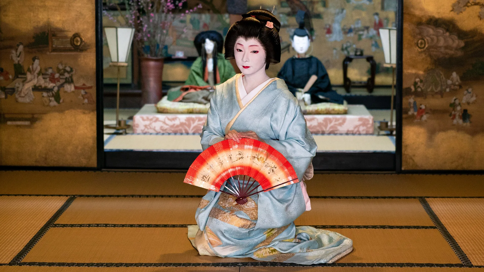 Geiko v bleděmodrém kimonu klečí na tatami před zástěnou se zlatými listy a drží před sebou červeno-bílý skládací vějíř; ilustrační fotografie tance geiko, nikoli tohoto představení